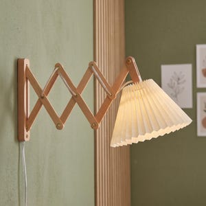 Puede incluir: Una lámpara de pared con un brazo extensible de madera clara estilo acordeón y una pantalla plisada en forma de cono. El accesorio incluye un cable de alimentación blanco que sale de la base, proporcionando una solución de iluminación funcional.