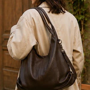 May include: A dark brown hobo bag with a slouchy silhouette, featuring a wide shoulder strap, a top zipper closure, and a metal hardware ring attachment on the side. The bag has a textured surface and is labeled with the text DARK BROWN.