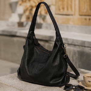 May include: A black hobo-style shoulder bag with a textured surface, a single top handle, and a detachable shoulder strap attached with gold-tone metal hardware. The bag features a zippered main compartment and a slouchy, unstructured silhouette.