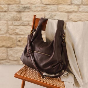 May include: A dark brown hobo bag with a pebbled texture, featuring a horizontal zippered pocket on the front, a wide shoulder strap, and antique brass-toned metal hardware clips attached to the sides of the bag for an adjustable strap.