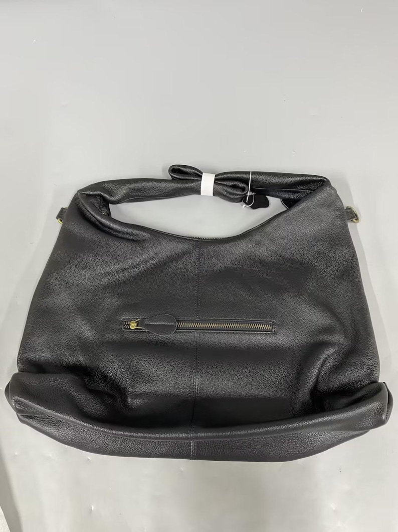 May include: A black hobo-style shoulder bag with a textured surface, featuring a single wide strap, a horizontal zippered pocket on the front with a matching black pull tab, and a small white tag attached to the top handle hardware.