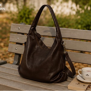 May include: A dark brown hobo bag with a slouchy silhouette, a single top handle, and a detachable shoulder strap with brass-toned hardware. The bag features a zippered top closure and a textured surface. The text "DARK BROWN" is visible in the corner.