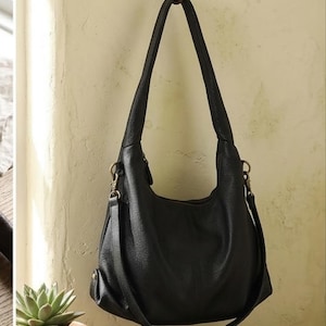 May include: A black hobo-style shoulder bag with a textured surface, featuring a single wide top handle and a detachable crossbody strap attached with brass-toned metal hardware. The bag has a slouchy, rounded silhouette and a zippered main closure.