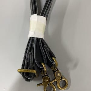 May include: A black strap folded into a loop and secured with a white paper band. The strap features two antique brass-toned metal swivel lobster claw clasps at one end and a rectangular brass-toned metal adjustment buckle along the length.
