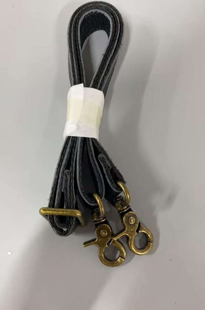 May include: A black strap folded into a loop and secured with a white paper band. The strap features two antique brass-toned metal swivel lobster claw clasps at one end and a rectangular brass-toned metal adjustment buckle along the length.