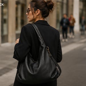 May include: A black hobo-style shoulder bag with a smooth texture, featuring a wide strap, a metallic ring attachment, and a zipper closure. The word BLACK is printed in the upper left corner of the image, indicating the color of the bag.