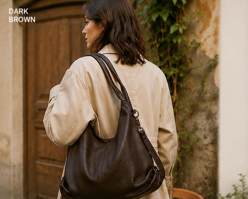 May include: A dark brown hobo bag with a slouchy silhouette, featuring a wide shoulder strap, a top zipper closure, and a metal hardware ring attachment on the side. The bag has a textured surface and is labeled with the text DARK BROWN.
