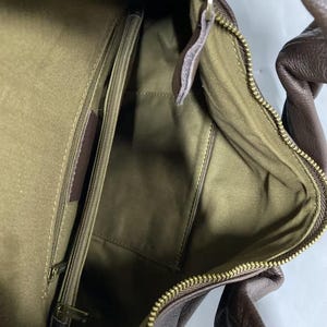 May include: A brown leather handbag interior featuring an olive green fabric lining, a central zippered compartment, and a gold-toned metal zipper closure. The bag has a textured exterior surface and a matching brown leather handle visible at the top.