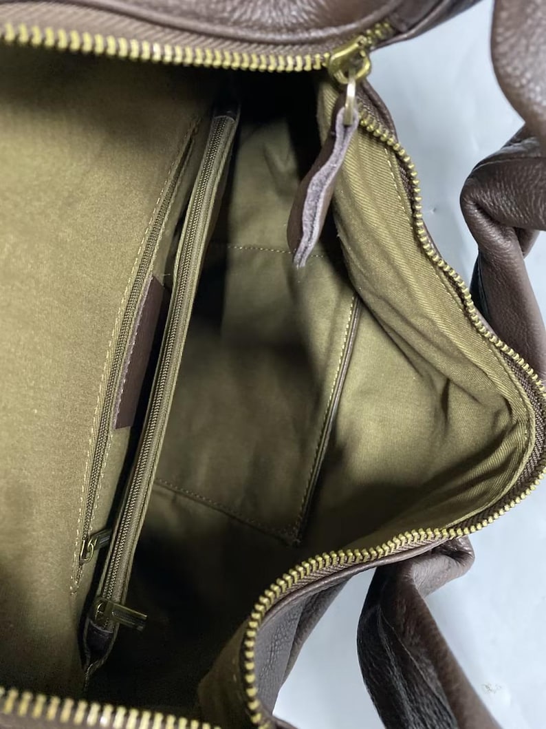 May include: A brown leather handbag interior featuring an olive green fabric lining, a central zippered compartment, and a gold-toned metal zipper closure. The bag has a textured exterior surface and a matching brown leather handle visible at the top.