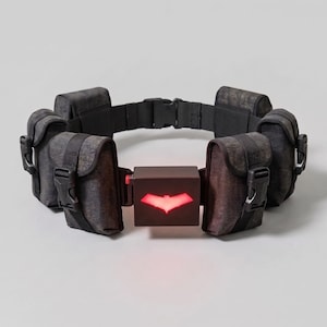 Peut inclure: Une ceinture utilitaire noire dotée de plusieurs pochettes avec des sangles en nylon noir et des boucles en plastique. Le centre de la ceinture comprend un boîtier noir rectangulaire avec un emblème rouge lumineux en forme de chauve-souris.