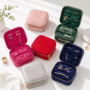 May include: A collection of square velvet jewelry boxes in pink, red, dark green, navy blue, and light gray. Each box features a gold-tone zipper, a soft interior lining, and dedicated compartments for rings, earrings, and necklaces.