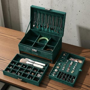 May include: A dark green velvet jewelry box with a hinged lid, featuring a necklace rack, multiple compartments, and two removable trays. The interior is lined with matching green fabric, containing various rings, bracelets, and two makeup brushes.