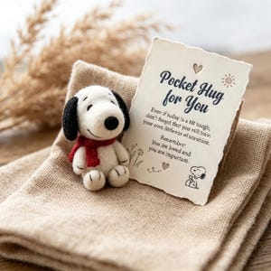 Può includere: Una piccola statuetta di cane in feltro bianco con orecchie nere e una sciarpa rossa accanto a un cartoncino rettangolare dai bordi irregolari. Il cartoncino riporta il testo Pocket Hug for You, un cuore, un sole e un disegno del cane.