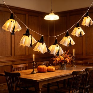 May include: A string of seven illuminated ghost decorations hanging in a row. Each ghost is draped in white fabric, wearing a small black pointed witch hat, and holding a miniature black broomstick. The ghosts emit a warm, soft yellow glow.