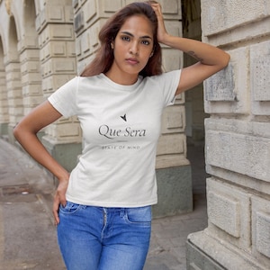 Op de afbeelding: Een wit T-shirt met korte mouwen voorzien van de zwarte tekst Que Sera en de zin STATE OF MIND op de borst, gecombineerd met een blauwe spijkerbroek. Het shirt heeft een ronde hals en een eenvoudig ontwerp met een klein zwart pijlicoon.