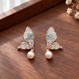 May include: A pair of gold-tone butterfly-shaped earrings featuring wings filled with light blue and white crushed stone fragments, accented with clear pear-shaped crystals and a single dangling white pearl on each earring.