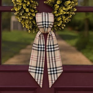 May include: A circular wreath composed of dense, small green leaves, featuring a long, hanging fabric ribbon tied in a knot at the centre. The ribbon displays a beige, black, white, and thin red plaid pattern with pointed ends.