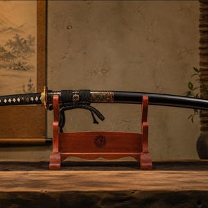 Peut inclure: Un sabre katana avec un fourreau laqué noir et une poignée enveloppée de cordon noir avec des accents blancs en forme de losange. Le sabre repose horizontalement sur un support de présentation en bois brun rougeâtre, avec une garde dorée.