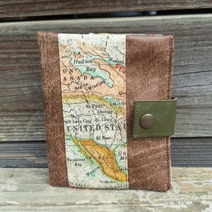 Può includere: Un porta passaporto in pelle marrone con un disegno a mappa. Il porta passaporto presenta una striscia di tessuto con una mappa vintage del Nord America. Ha un cinturino verde oliva con una chiusura a scatto in bronzo. Il porta passaporto è su una superficie di legno.