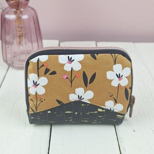 Può includere: Una piccola pochette con cerniera caratterizzata da una sezione superiore giallo senape con un motivo floreale bianco e foglie nere, contrastata da una sezione inferiore nera con un design intrecciato testurizzato. La pochette ha una cerniera scura.