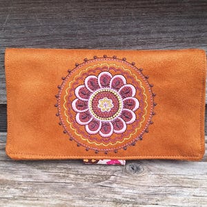 May include: A burnt orange clutch with an embroidered floral design in shades of burgundy, pink, and gold. The clutch is rectangular and made of a suede-like material, with a small patterned tab closure.