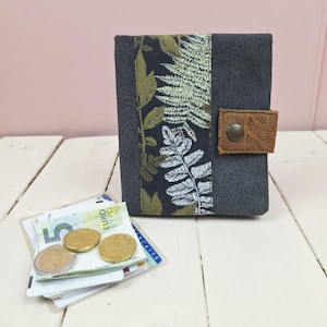 Op de afbeelding: Een grijze vilten portemonnee met een decoratief paneel met een botanische print in groen en wit. De portemonnee heeft een bruine leren tabsluiting met een metalen drukknop. Euro valuta en munten zijn zichtbaar.