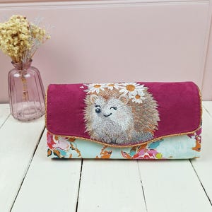 Op de afbeelding: Een bordeaux clutch met een bloemenprint basis en een flap met een geborduurde egel met een madeliefjeskroon. De clutch heeft een gouden rand en wordt gepresenteerd op een wit houten oppervlak.