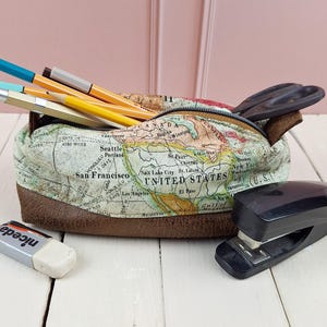 May include: A fabric pencil case with a map design, filled with pens, pencils, and scissors. The case has a brown leather bottom and a zipper closure. A stapler and an eraser are next to the case.