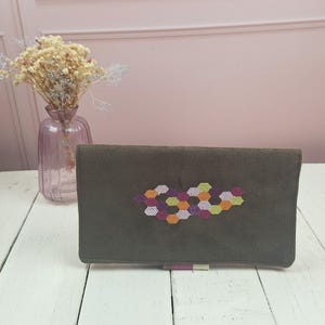 Può includere: Un portafoglio rettangolare in camoscio marrone scuro con un motivo centrale ricamato di piccoli esagoni multicolori nelle tonalità dell'arancione, viola, rosa e giallo chiaro. Il portafoglio ha una consistenza liscia e opaca.