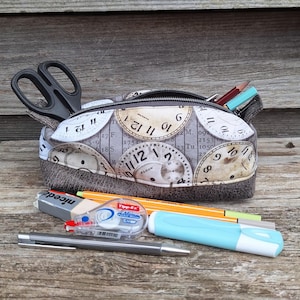 Puede incluir: Estuche de tela con un diseño de esfera de reloj, abierto para revelar tijeras y lápices. Otros artículos de papelería, como un bolígrafo, una goma de borrar y un resaltador, están colocados delante. El estuche mide unos 20 cm de largo.