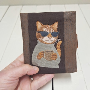 Può includere: Un portafoglio marrone con un pannello centrale in tessuto marrone scuro che presenta il ricamo di un gatto tigrato arancione con occhiali da sole blu scuro e un maglione grigio, mentre tiene in mano una tazza marrone. I lati sono in similpelle testurizzata.