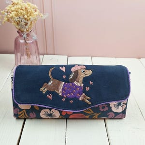 Könnte beinhalten: Eine rechteckige marineblaue Stofftasche mit einem gestickten braunen Dackel, der einen violetten Pullover und ein rosa Barett trägt, umgeben von kleinen rosa Herzen. Der untere Teil der Tasche zeigt ein Blumenmuster mit rosa und orangefarbenen Blüten.