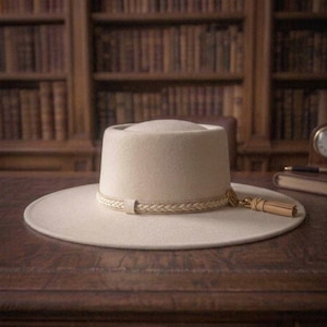 May include: A cream-coloured wide-brim hat with a flat crown. The hat features a braided band around the base, accented with a small circular emblem and a matching leather tassel hanging from the side of the crown, resting on the wide brim.