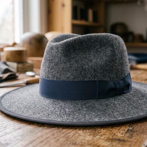 Può includere: Un cappello fedora in feltro grigio antracite con tesa larga, bordo rifinito in grigio scuro e un nastro in gros-grain blu navy che circonda la corona. Il cappello presenta un design classico con corona pizzicata e una texture uniforme.