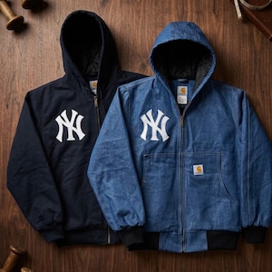 Pode incluir: Duas jaquetas com capuz e zíper, apresentando o logotipo branco dos New York Yankees no peito. Uma jaqueta é azul-marinho sólida e a outra é de tecido jeans azul. Ambas possuem punhos pretos canelados, forro interno preto e uma etiqueta Carhartt.