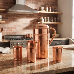 May include: A polished copper water pitcher with a matching lid and handle, accompanied by three matching copper tumblers. The pitcher features a textured band around the center, and the tumblers have a similar horizontal line pattern.