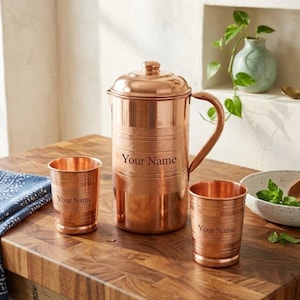 May include: A polished copper pitcher with a lid and handle, accompanied by two matching copper tumblers. Each piece features horizontal etched bands and the custom text Your Name engraved on the front surface of the pitcher and both cups.