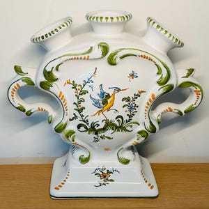 Pode incluir: Um vaso de cerâmica branca com três aberturas superiores e duas alças laterais. A superfície apresenta volutas verdes e laranjas pintadas à mão, motivos florais e uma ilustração central de um pássaro amarelo e azul em pleno voo.