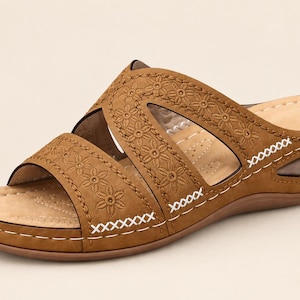 May include: A tan slide sandal featuring a wedge heel and two wide straps across the top. The straps are decorated with an embossed floral pattern and accented with white cross-stitch detailing along the edges. The footbed is a light beige color.