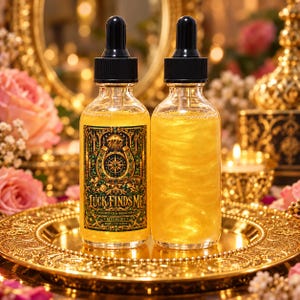 May include: Two clear glass dropper bottles filled with shimmering gold liquid. One bottle features a label with green and gold ornate details, a horseshoe, and the text Luck Finds Me, Good Luck & Blessings, Ritual Oil, 60ml, 2.02 fl oz.