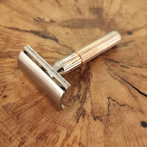 Puede incluir: Una maquinilla de afeitar de seguridad vintage con cabezal plateado y mango dorado. La maquinilla es de metal y está diseñada para afeitarse. La maquinilla está sobre una superficie de madera.