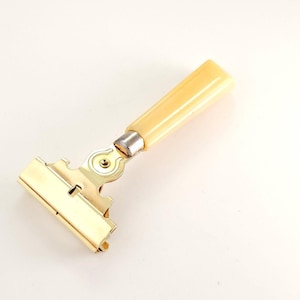 May include: A vintage metal and cream-coloured paper clip with a hinged design. The clip has a rectangular shape with a gold finish, and a cream-coloured handle. The clip is designed to hold papers together.
