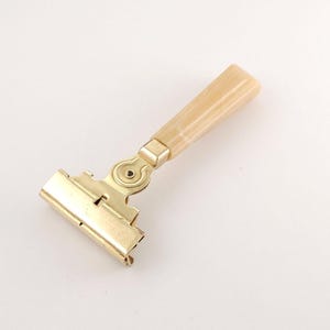 Könnte beinhalten: Ein goldfarbener Sicherheitsrasierer aus Metall mit einem klappbaren Kopfmechanismus und einem glatten, cremefarbenen Griff in Marmoroptik. Der Rasierer hat eine metallische Oberfläche und einen markanten Drehpunkt zwischen Kopf und Griff.