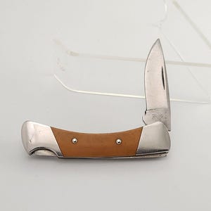 Op de afbeelding: Een gedeeltelijk opengeklapt zakmes met een bruin handvat en een zilverkleurig lemmet. Het lemmet is gemarkeerd met het woord "Buck". Het mes ligt op een witte ondergrond.