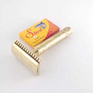 May include: A gold-toned metal safety razor with a textured handle and a comb-style head, positioned next to a small yellow and red rectangular box labeled Shark Super Stainless Blades with a blue shark graphic and the number 5.
