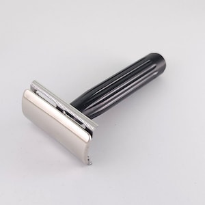 May include: A safety razor featuring a polished silver-tone metal head with a comb-style blade guard and a black handle with vertical ridges. The handle is cylindrical and tapers slightly toward the end, connecting to the metallic razor head.