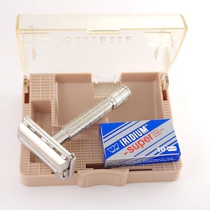 Puede incluir: Una maquinilla de afeitar de seguridad cromada vintage con un mango texturizado descansa en un estuche de plástico beige abierto, junto a una caja azul y blanca de cuchillas Iridium Super. La maquinilla y las cuchillas son visibles.