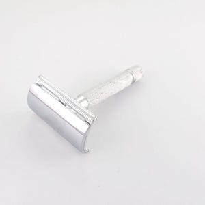 May include: A silver-toned safety razor featuring a polished, smooth head and a textured, knurled handle designed for a secure grip. The metal construction has a reflective finish, and the head assembly includes a comb-like guard along the edge.