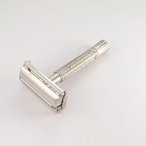 Puede incluir: Una maquinilla de afeitar de seguridad plateada vintage con un mango texturizado y un cabezal giratorio. La maquinilla está diseñada para afeitarse y presenta un diseño clásico. El metal tiene un aspecto ligeramente desgastado, lo que sugiere edad y uso.
