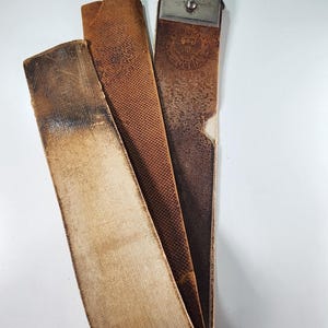 Puede incluir: Un cuero antiguo para afilar navajas. El cuero está hecho de varias capas de cuero en diferentes tonos marrones, con un cierre de metal en la parte superior. El cuero muestra signos de desgaste y envejecimiento.